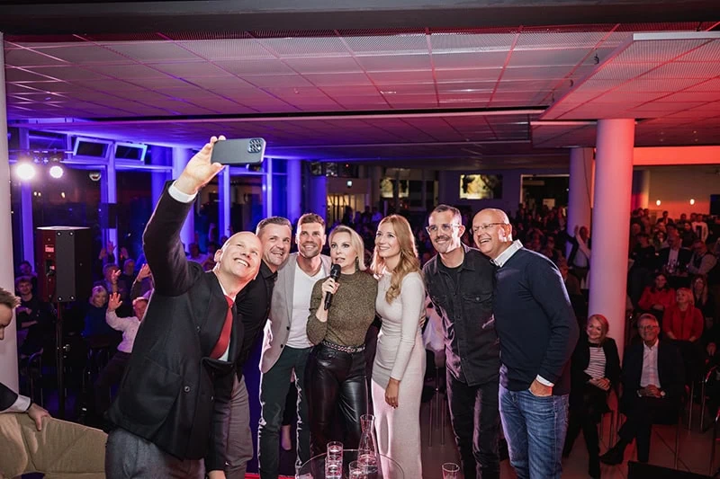 Sieben Personen posieren für ein Selfie in einer Veranstaltungshalle mit bunten Lichtern.