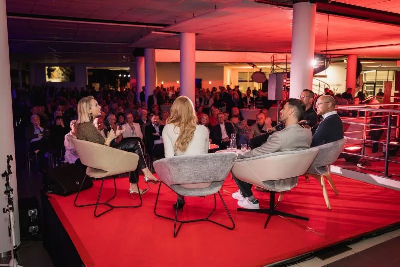 Vier Personen sitzen auf einem Podium während einer Talkrunde mit Publikum im Hintergrund.